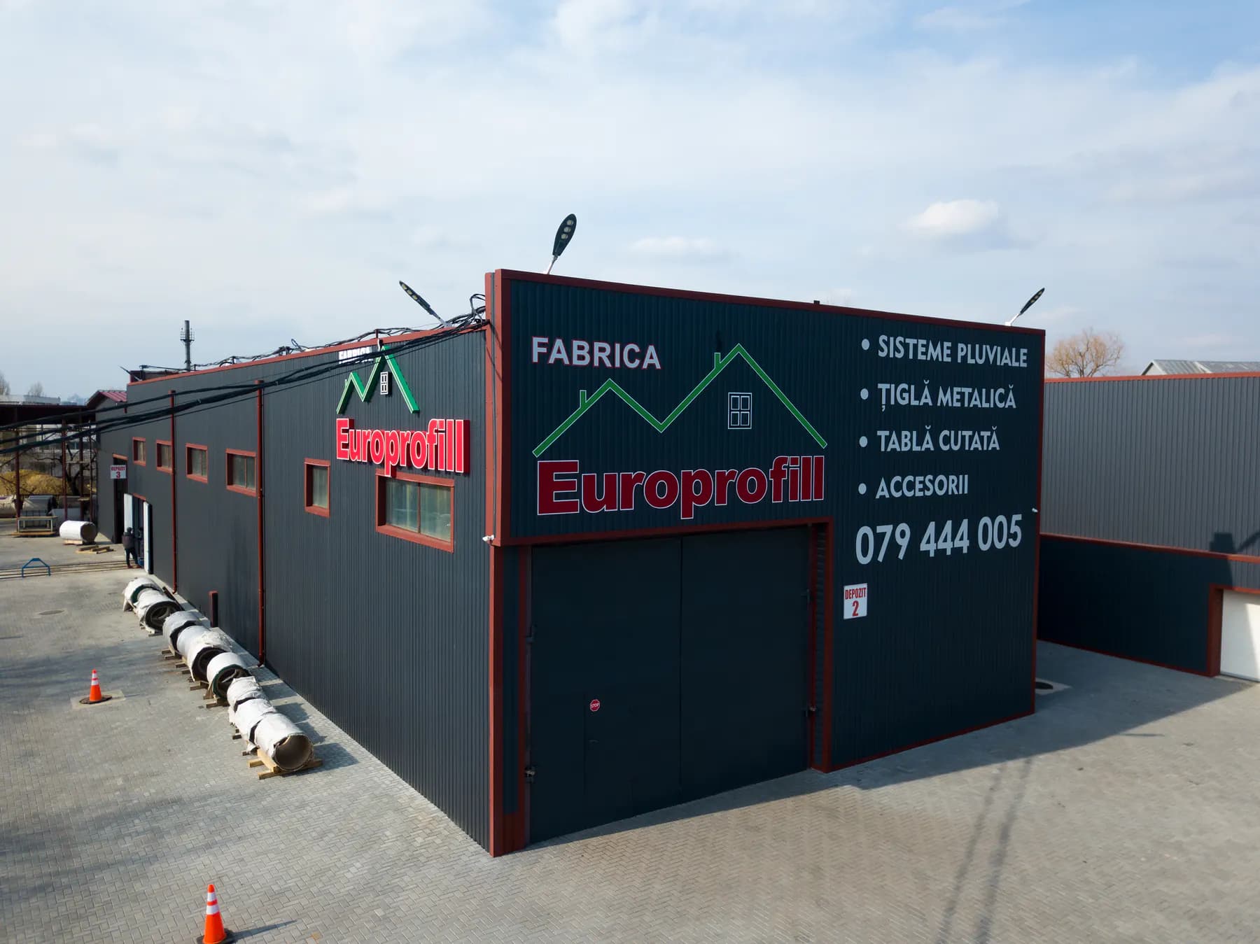 Vedere aeriană a clădirii fabricii Europrofill, gri închis cu borduri roșii și inscripția „Fabrica Europrofill”, cu rulouri de oțel aliniate în curte