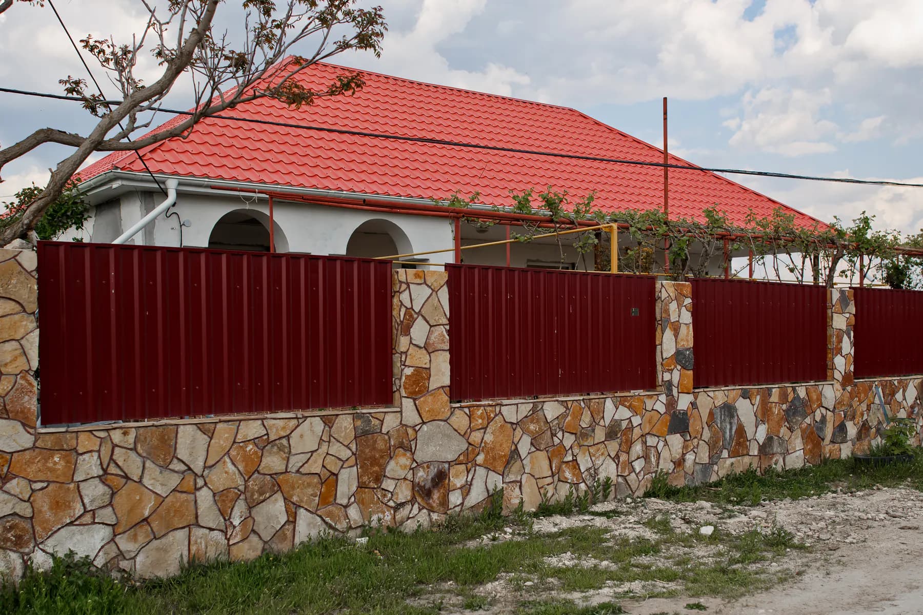 Gard cu panouri din tablă profilată vișinie între stâlpi din piatră naturală, în fața unei case cu acoperiș din țiglă metalică roșie
