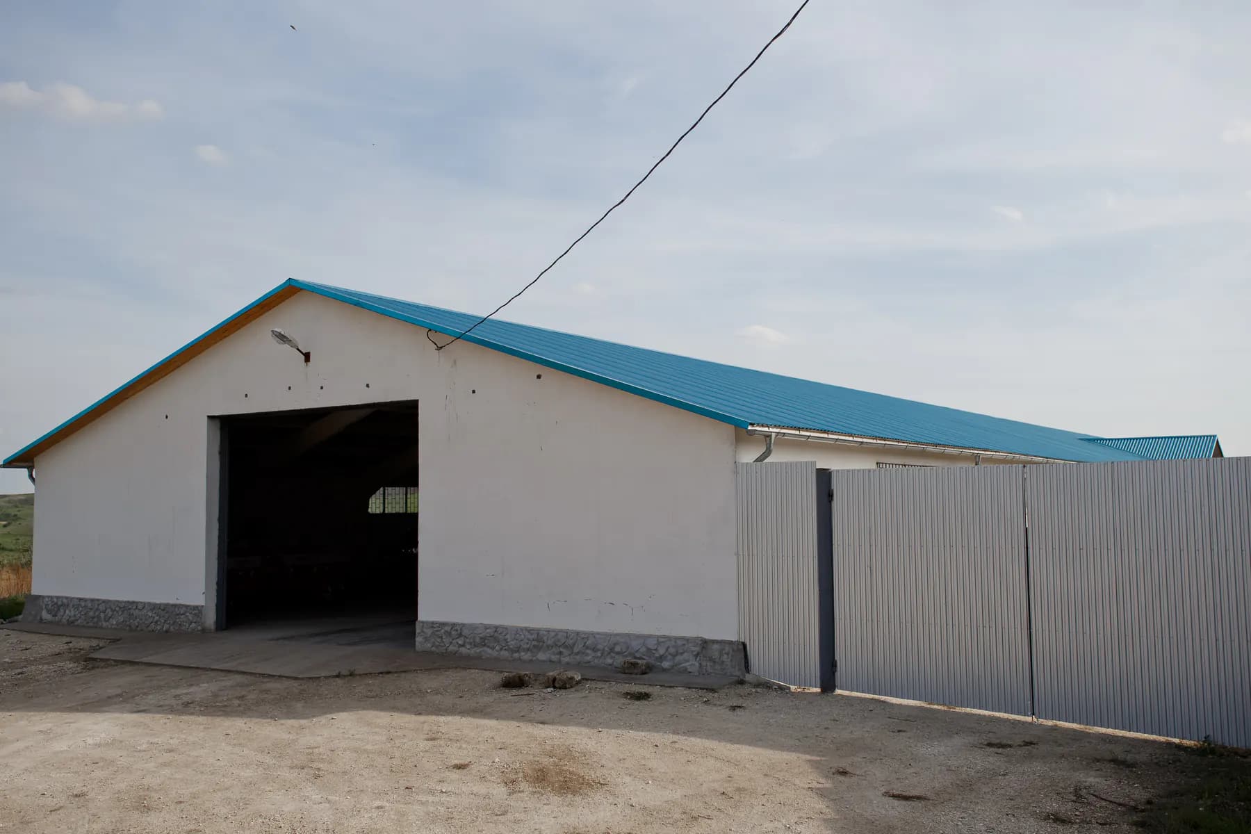Hală mare cu un nivel, cu acoperiș în două ape din tablă profilată albastru-turcoaz și intrare largă, lângă un gard din tablă profilată gri deschis
