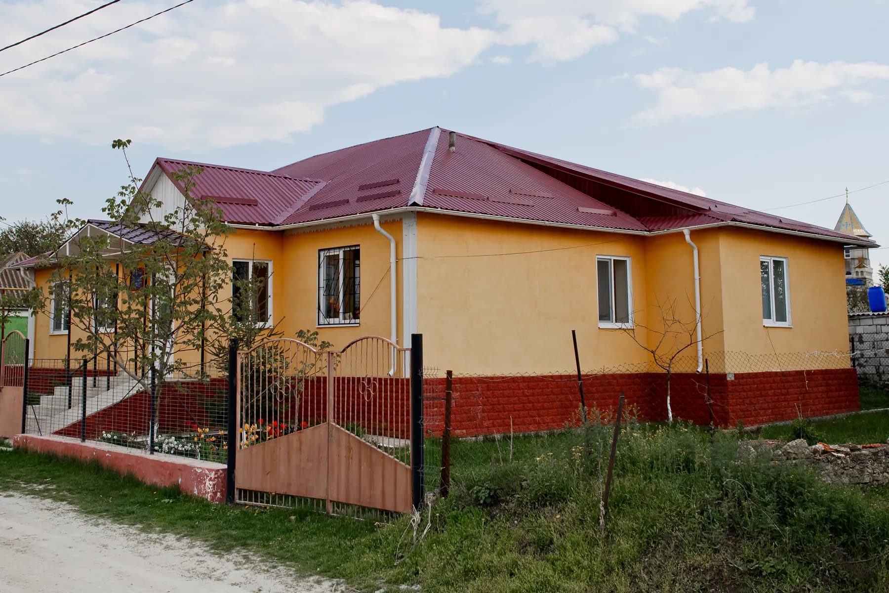 Casă galbenă cu un nivel, cu acoperiș din tablă profilată vișinie, burlane albe și soclu roșu