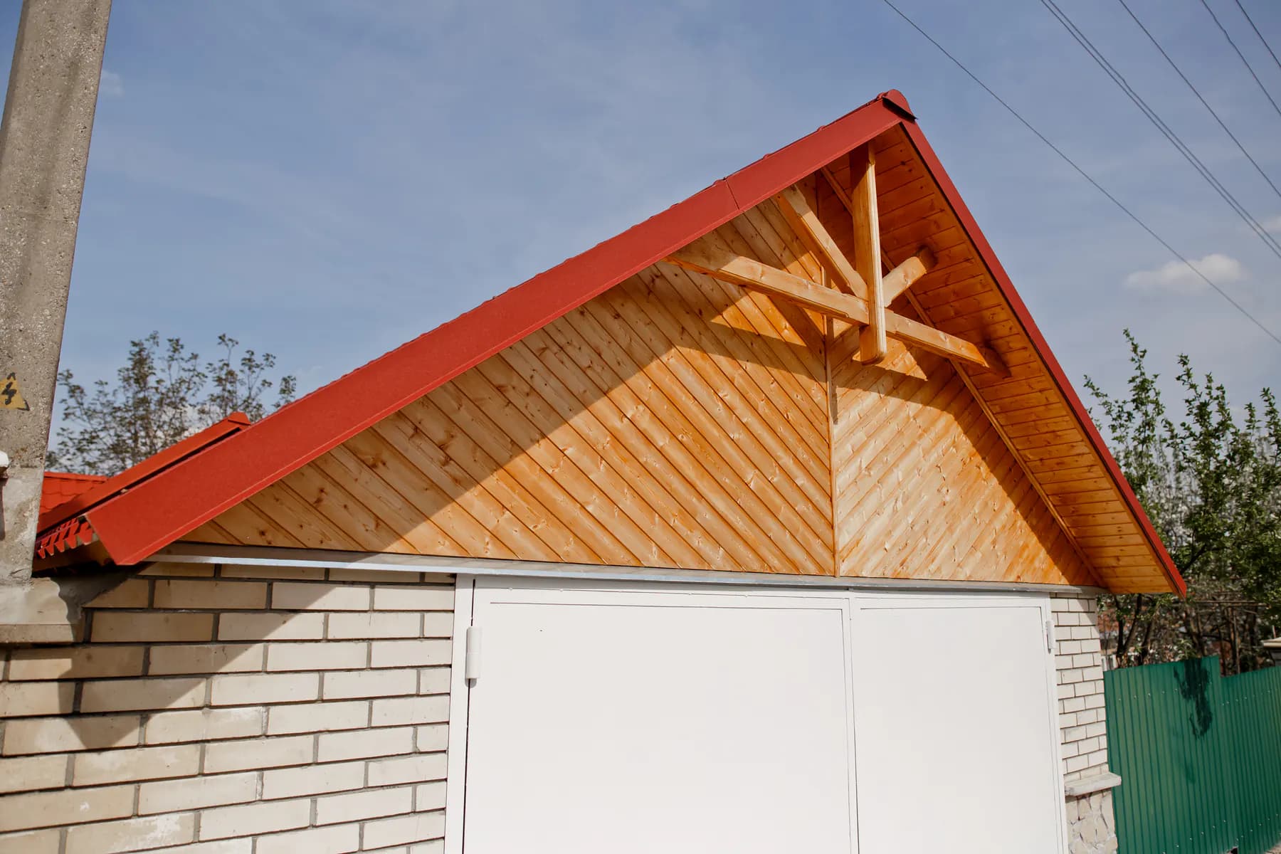 Garaj cu fronton placat cu lambriu de lemn și grindă decorativă, sub un acoperiș metalic roșu; porți albe dedesubt