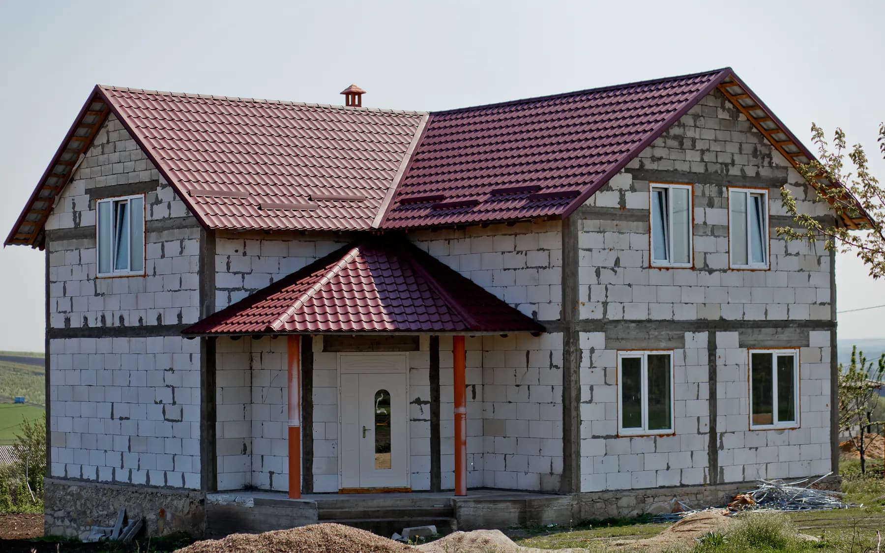 Casă cu două niveluri în construcție, cu zidărie netencuită și acoperiș din țiglă metalică vișinie, inclusiv peste copertina intrării