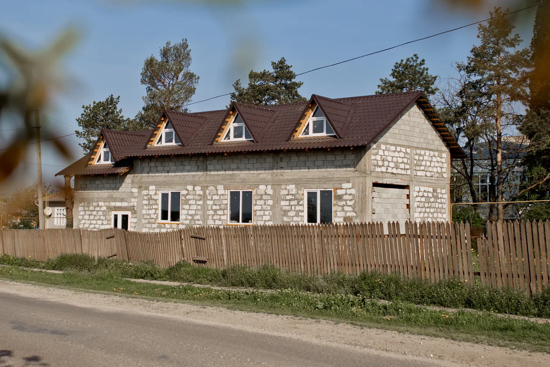 Casă cu două niveluri în construcție, cu mai multe lucarne și acoperiș din țiglă metalică maro închis; pini în spate și gard din șipci de lemn