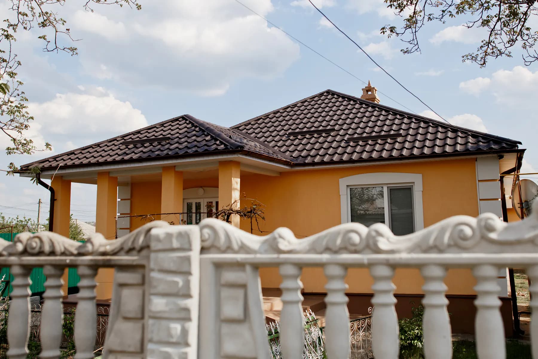 Casă cu fațadă portocalie și coloane, acoperită cu țiglă metalică maro închis, văzută peste un gard alb cu baluștri