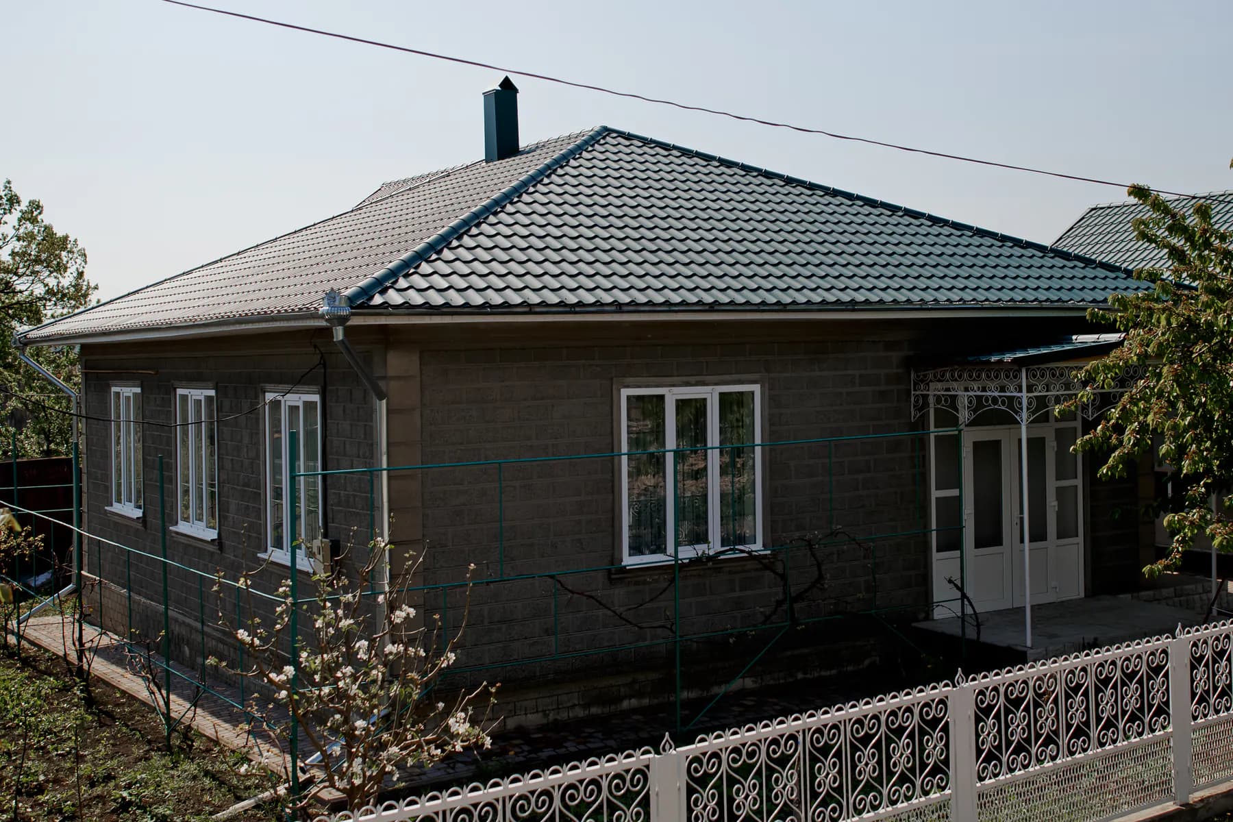 Casă cu un nivel din blocuri decorative gri închis, cu acoperiș în patru ape din țiglă metalică verde închis și gard alb ornamental