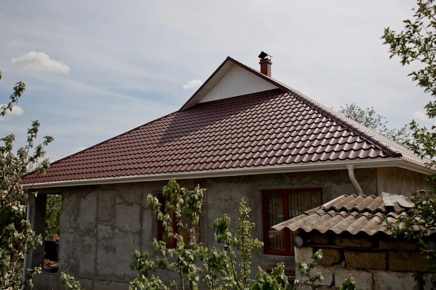 Acoperiș din țiglă metalică maro-roșcat, cu un fronton mic placat în alb și coș din cărămidă, văzut printre ramuri